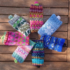 May include: Seven colorful knitted phone cases with different patterns. The cases are made of wool and have a pom-pom on top. The cases are arranged in a circle on a wooden surface.
