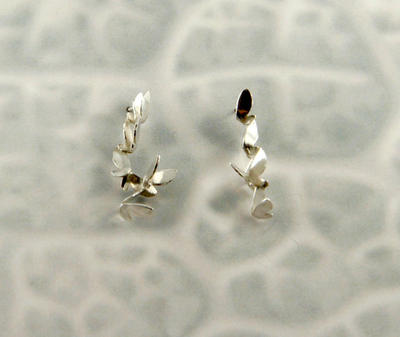 May include: A pair of silver earrings with a delicate, leaf-like design. Each earring features multiple small, curved leaves that are connected to form a flowing, organic shape.
