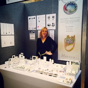 May include: A woman stands behind a table displaying handmade jewelry. The jewelry is made of silver and features small sculptures of buildings. The sign behind her reads "Agnieszka Maksymiuk creates jewelry which are small sculptures that become wearable pieces of art. Handmade Precious Jewelry. MAKSYMI"