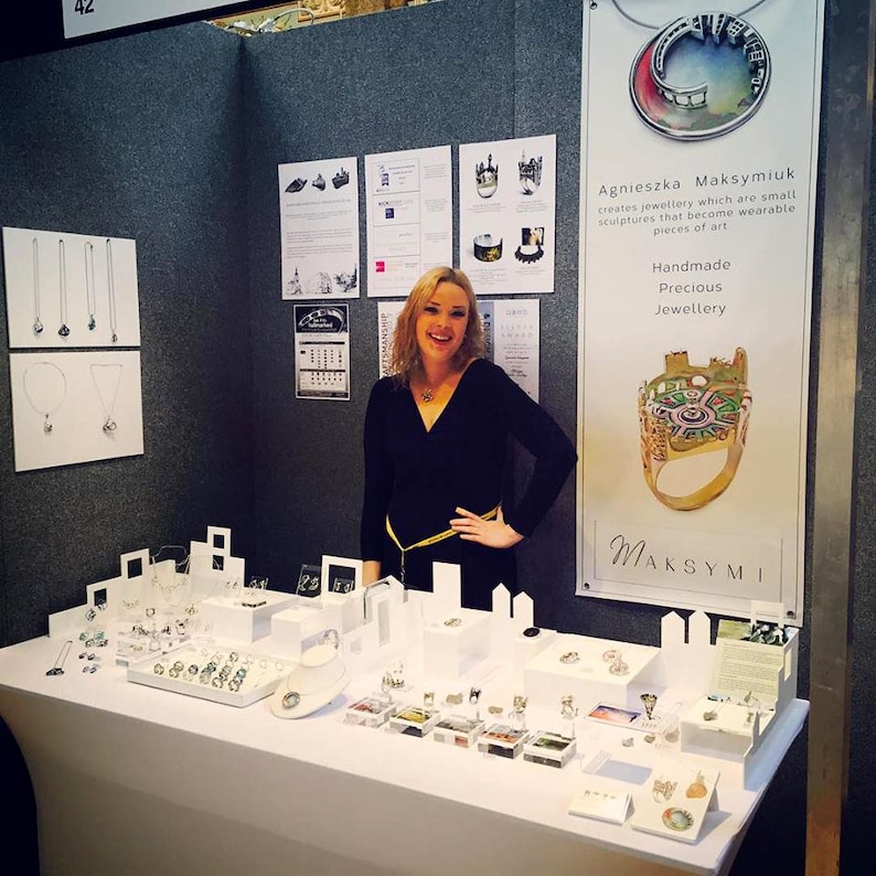 May include: A woman stands behind a table displaying handmade jewelry. The jewelry is made of silver and features small sculptures of buildings. The sign behind her reads "Agnieszka Maksymiuk creates jewelry which are small sculptures that become wearable pieces of art. Handmade Precious Jewelry. MAKSYMI"