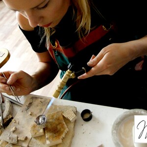 May include: A person uses a torch to heat a piece of metal jewelry while holding it with tweezers. The metal is sitting on a ceramic tile with small holes. There are other tools and materials on the table, including a small bowl, a pair of pliers, and a pencil.
