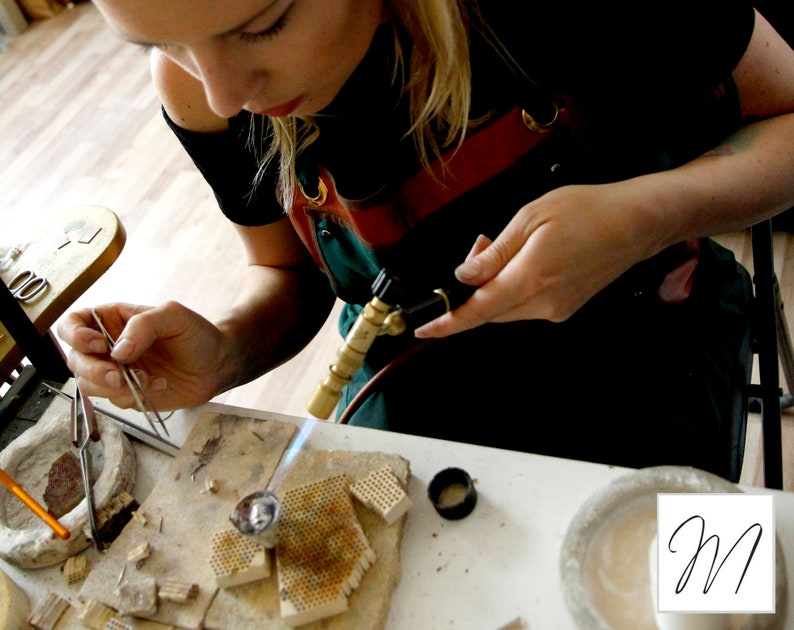 May include: A person uses a torch to heat a piece of metal jewelry while holding it with tweezers. The metal is sitting on a ceramic tile with small holes. There are other tools and materials on the table, including a small bowl, a pair of pliers, and a pencil.
