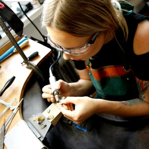 May include: A person wearing safety glasses and a green and orange apron is working on a piece of jewelry with a small tool. The person is sitting at a workbench with a variety of tools and materials.