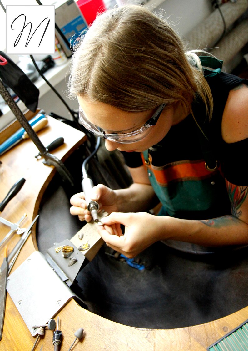 May include: A person wearing safety glasses and a green and orange apron is working on a piece of jewelry with a small tool. The person is sitting at a workbench with a variety of tools and materials.