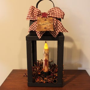 May include: Black rectangular lantern with a handle, a battery-operated candle, and a decorative bow. The bow is red and white checkered. The lantern has a tag that reads "Primitives". The candle has a red star design.