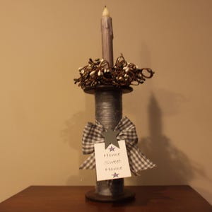May include: A decorative candle holder made from a large wooden spool, painted gray. It features a bow, a tag that says "Home Sweet Home", and a candle. The top is decorated with small white berries and brown twigs.