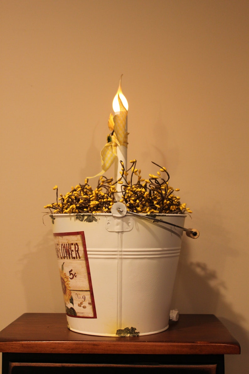 May include: A white metal bucket with a sunflower design and a decorative candle. The bucket is filled with yellow berries and has a handle. The candle has a yellow and white patterned design.