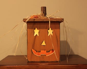 May include: A wooden Halloween decoration shaped like a jack-o'-lantern. The boxy structure has star-shaped eyes, a triangle nose, and a carved orange mouth. The top has a wooden stem and straw accents.