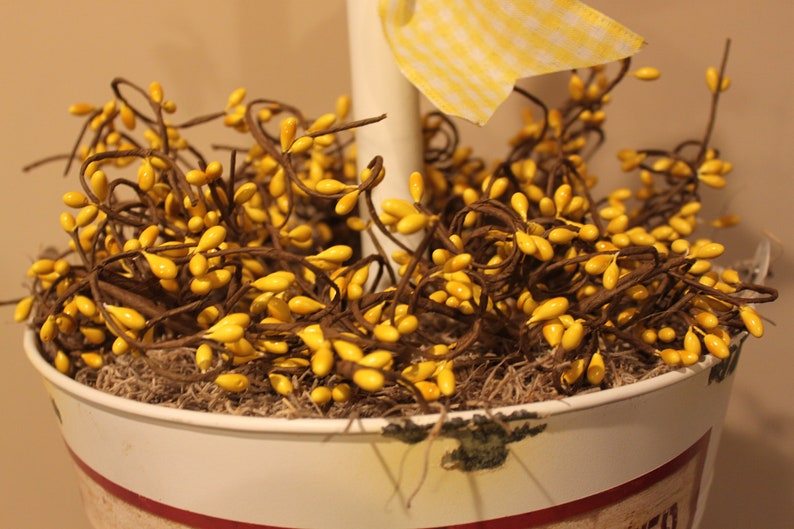 May include: A decorative arrangement featuring yellow berry stems in a white pot with a red stripe. The pot is filled with moss and a yellow gingham ribbon. The berries are a bright, cheerful yellow.