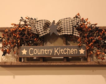 May include: A wooden wall sign featuring the text Country Kitchen in light lettering, flanked by two stars. The sign is decorated with a black and white checkered fabric bow, a dark gray star cutout, and two clusters of orange and black pip berries.