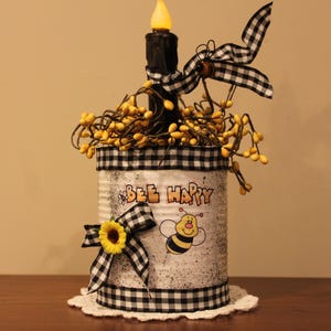 May include: A decorative tin can with a black and white checkered pattern, featuring the words "BEE HAPPY" and a cartoon bee. It is adorned with a sunflower, bows, and yellow berries, topped with a black candle.