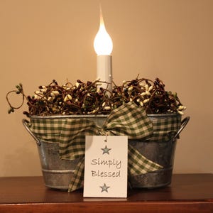 May include: A galvanized metal oval basin filled with dark brown and cream-colored pip berries, featuring a central electric candle with a flame-shaped bulb. A green and white gingham ribbon is tied around the basin with a white tag reading Simply Blessed.