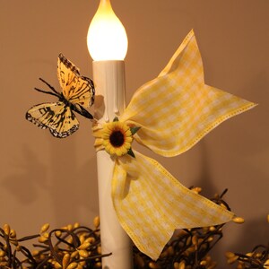 May include: A decorative candle with a warm, glowing flame. It is adorned with a yellow and white gingham bow, a sunflower, and a butterfly with black and yellow markings. The candle sits atop a bed of yellow berries.