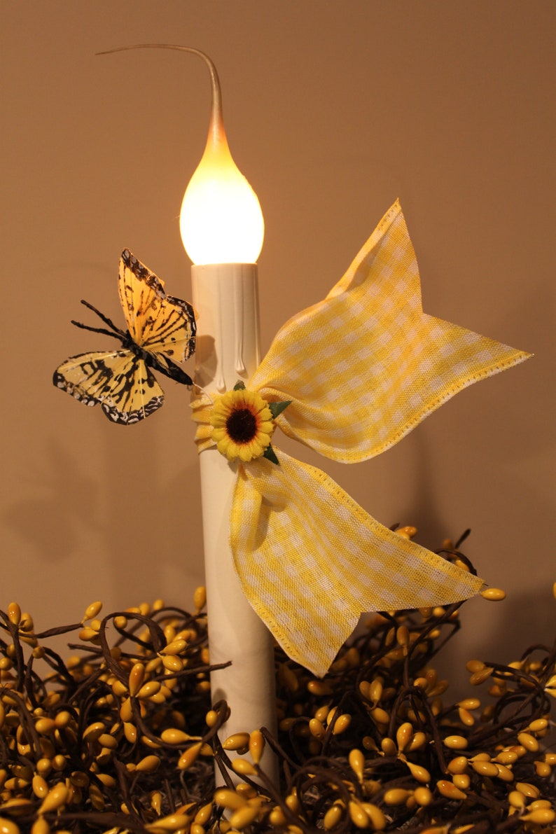May include: A decorative candle with a warm, glowing flame. It is adorned with a yellow and white gingham bow, a sunflower, and a butterfly with black and yellow markings. The candle sits atop a bed of yellow berries.