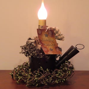 May include: A black metal candle holder shaped like a small watering can, featuring a lit flame-shaped bulb. It is decorated with a green berry wreath and a rectangular tag with brown edges that reads Simply Blessed with two small blue star icons.