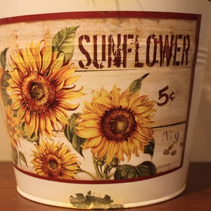 May include: A white metal bucket with a distressed finish, featuring a decorative panel with sunflowers and the word "SUNFLOWER". The panel also includes the number "5&cent;" and the text "No. 9".