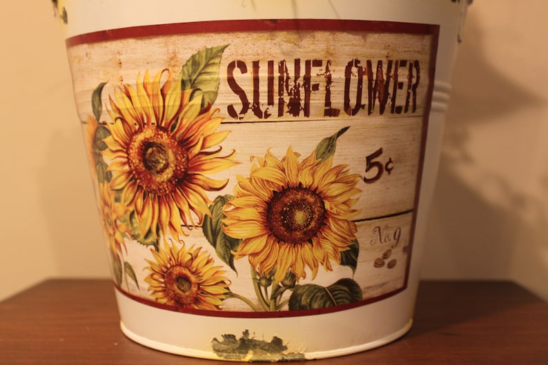 May include: A white metal bucket with a distressed finish, featuring a decorative panel with sunflowers and the word "SUNFLOWER". The panel also includes the number "5&cent;" and the text "No. 9".