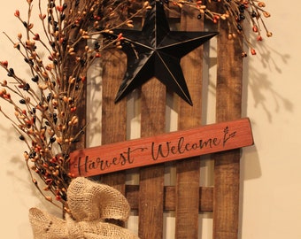 May include: A rustic wooden fence-style wall hanging with a black star and a burlap bow. The fence is adorned with a berry wreath and a sign that reads "Harvest Welcome". The color palette includes brown, black, red, and beige.