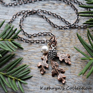 May include: A copper-toned necklace with an oak leaf pendant, a pearl, and a small pinecone charm. The necklace rests on a wooden surface, framed by green pine branches. The text "Kathryn's Collection" is visible.