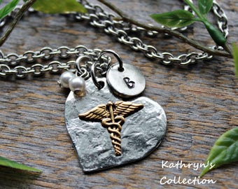 Puede incluir: Collar plateado con un colgante en forma de corazón con un símbolo médico dorado. El collar incluye un pequeño disco con la letra "B" y dos pequeñas perlas. La cadena es de estilo eslabón grueso, sobre un fondo de madera.
