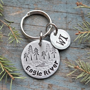 May include: A silver-toned metal keychain featuring two circular charms. The larger charm is stamped with a forest of pine trees, a loon, and the text Eagle River. The smaller charm is stamped with the letters WI.