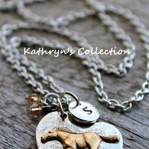 May include: Silver-toned necklace with a heart-shaped pendant featuring a gold-colored horse. The pendant has a hammered texture and includes a small disc with the letter "S". The necklace also has a small cluster of amber-colored beads. The text "Kathryn's Collection" is visible.