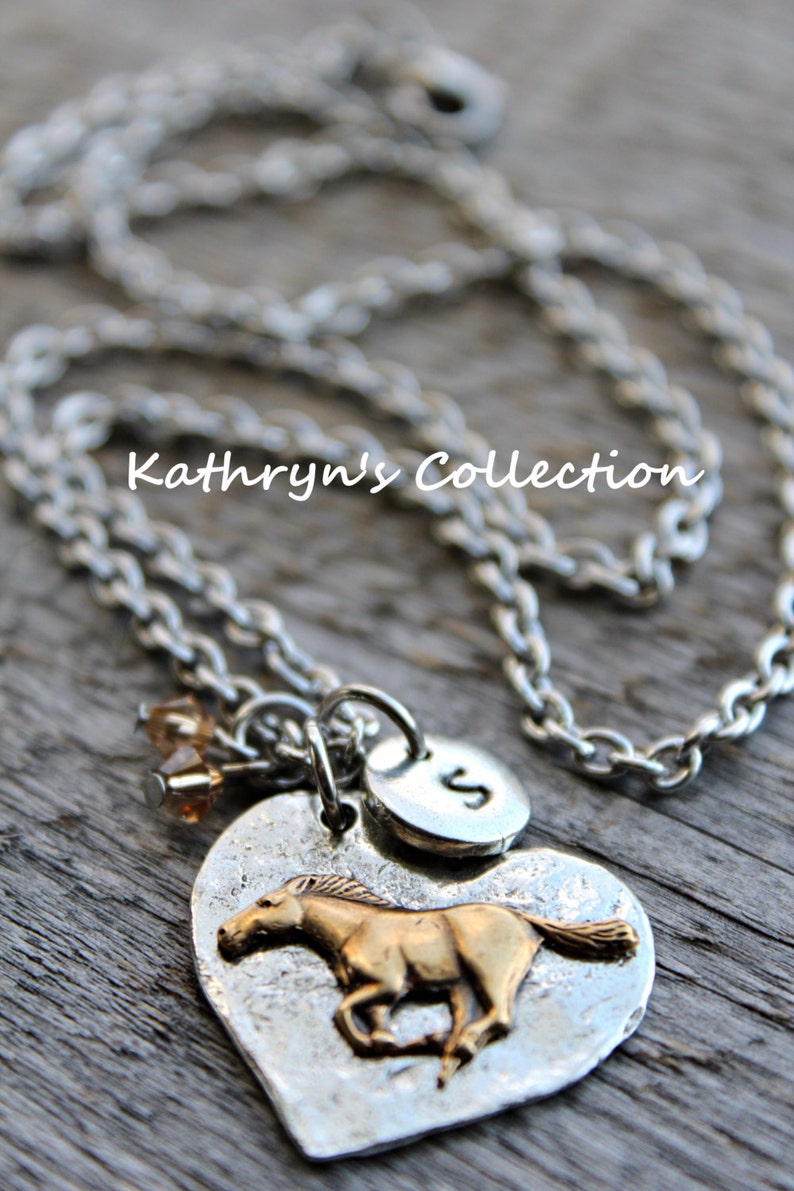 May include: Silver-toned necklace with a heart-shaped pendant featuring a gold-colored horse. The pendant has a hammered texture and includes a small disc with the letter "S". The necklace also has a small cluster of amber-colored beads. The text "Kathryn's Collection" is visible.