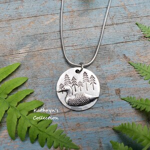 May include: A silver necklace with a round pendant featuring a loon and a forest scene. The pendant is textured and has a rustic look. The necklace is on a wooden surface with green ferns.