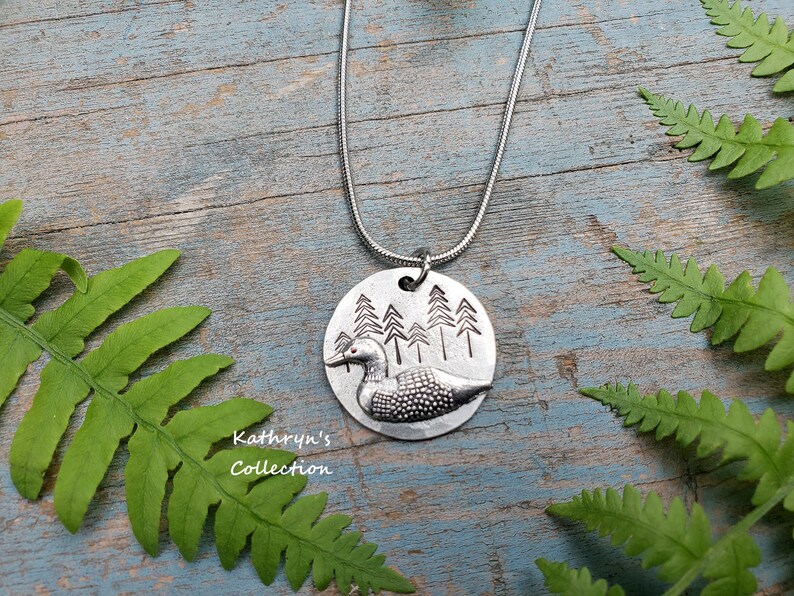 May include: A silver necklace with a round pendant featuring a loon and a forest scene. The pendant is textured and has a rustic look. The necklace is on a wooden surface with green ferns.
