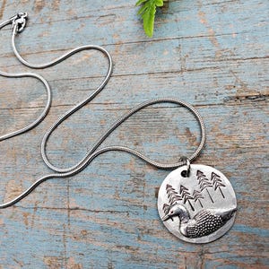 May include: A silver chain necklace with a round pendant featuring a loon in a forest setting. The pendant is made of metal and has a detailed design of a loon swimming in a lake with trees in the background.