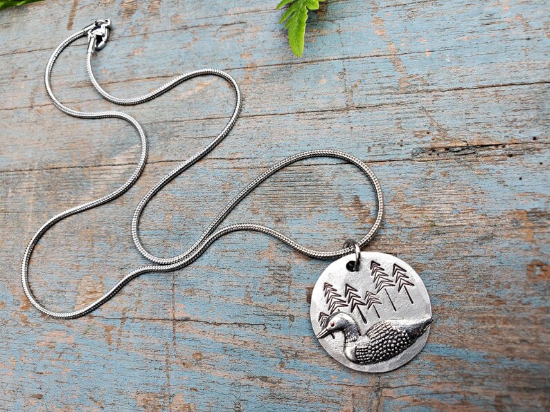 May include: A silver chain necklace with a round pendant featuring a loon in a forest setting. The pendant is made of metal and has a detailed design of a loon swimming in a lake with trees in the background.