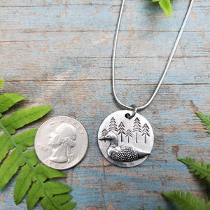 May include: A silver necklace with a round pendant featuring a loon in a forest scene. The pendant is about the size of a quarter.