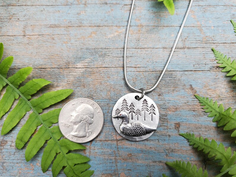 May include: A silver necklace with a round pendant featuring a loon in a forest scene. The pendant is about the size of a quarter.