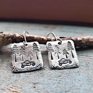 May include: A pair of silver earrings with a design of trees and a small car. The earrings are hanging from a branch.