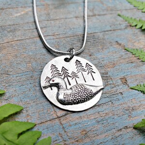 May include: A silver necklace with a round pendant featuring a loon in a forest setting. The loon is detailed with a textured body and red eyes. The pendant is attached to a silver chain.