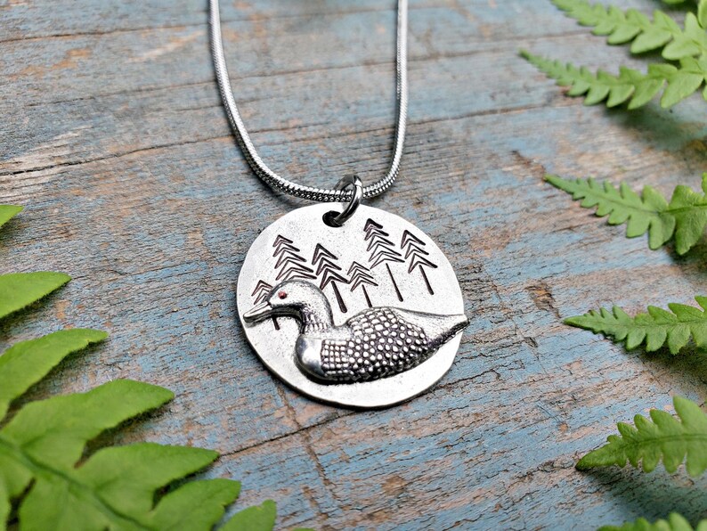 May include: A silver necklace with a round pendant featuring a loon in a forest setting. The loon is detailed with a textured body and red eyes. The pendant is attached to a silver chain.