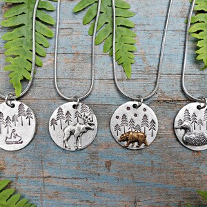 May include: Four silver necklaces with round pendants featuring different woodland scenes. The pendants depict a moose, a bear, a loon, and a person riding a snowmobile. Each pendant has a background of pine trees.