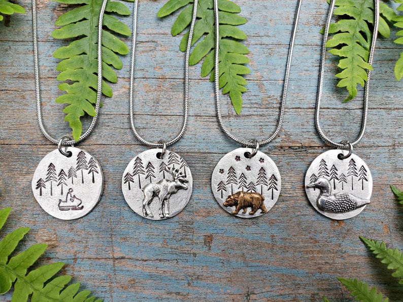 May include: Four silver necklaces with round pendants featuring different woodland scenes. The pendants depict a moose, a bear, a loon, and a person riding a snowmobile. Each pendant has a background of pine trees.