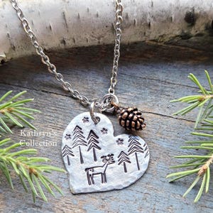 May include: Silver-toned necklace with a heart-shaped pendant featuring a forest scene with trees and a deer. A small pine cone charm hangs next to the pendant. The chain is a linked style.
