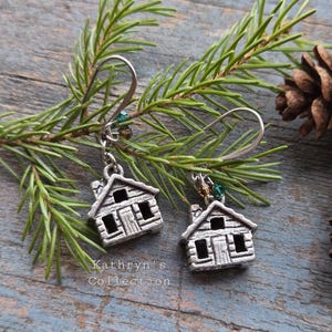 May include: Silver-toned cabin earrings with a rustic design, featuring a small chimney and windows. The earrings hang from silver hooks and are accented with green and brown beads. The earrings are displayed with pine needles and a pine cone.