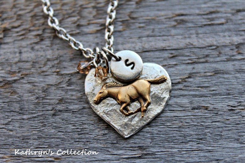 May include: Silver-toned necklace with a heart-shaped pendant featuring a running horse charm. The pendant has a textured surface and a small, round charm with the letter "S". Brown crystal beads dangle near the pendant. The text "Kathryn's Collection" is visible.