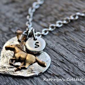 May include: Silver-toned necklace with a heart-shaped pendant featuring a textured surface and a gold-colored horse. A small, round charm with the letter "S" is attached. The necklace is on a wooden surface. Text at the bottom reads "Kathryn's Collection."
