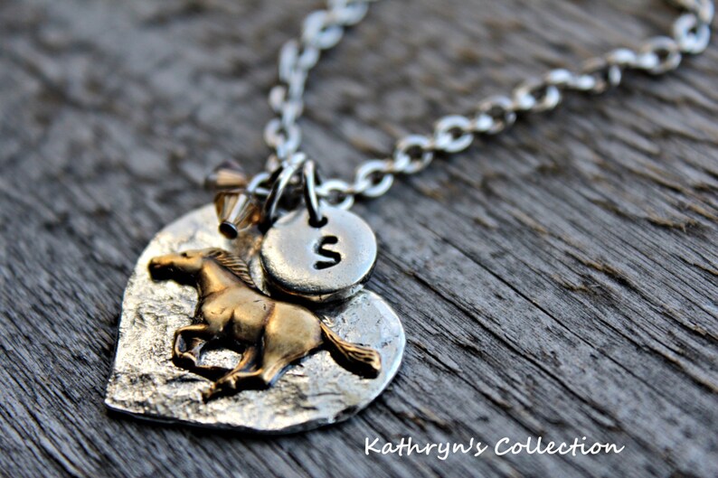 May include: Silver-toned necklace with a heart-shaped pendant featuring a textured surface and a gold-colored horse. A small, round charm with the letter "S" is attached. The necklace is on a wooden surface. Text at the bottom reads "Kathryn's Collection."