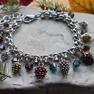 May include: A silver charm bracelet featuring pinecone and crystal accents. The bracelet has a chain link design with various sizes of pinecone charms and faceted crystal beads in shades of green, brown, and gold.