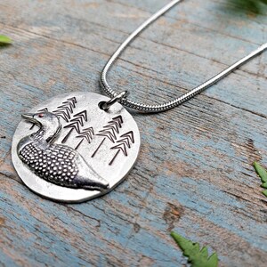 May include: A silver necklace with a round pendant featuring a loon and a design of stylized pine trees.