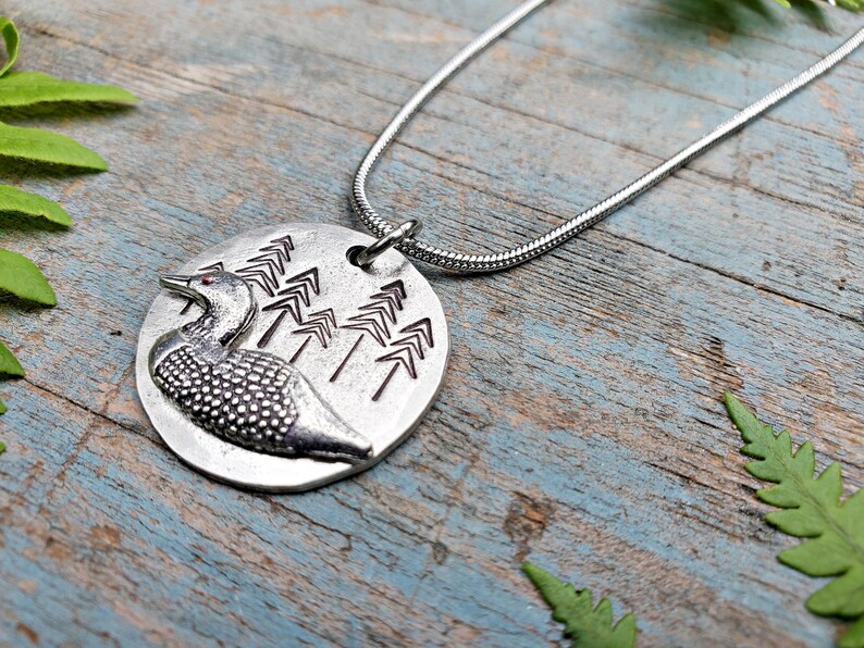 May include: A silver necklace with a round pendant featuring a loon and a design of stylized pine trees.