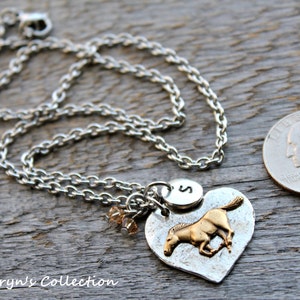 May include: Silver-toned necklace with a heart-shaped pendant featuring a gold-colored horse. The pendant includes an initial charm and small crystal beads. A quarter is next to the necklace for size reference. The necklace is on a wooden surface.