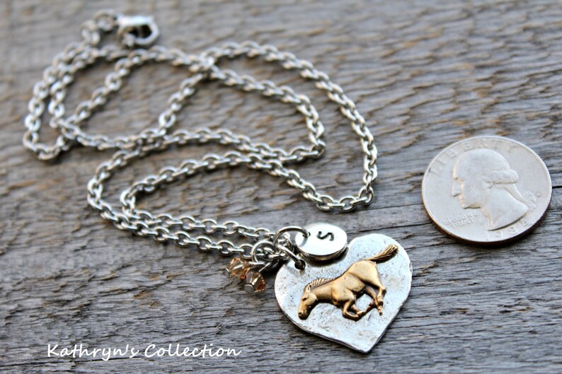 May include: Silver-toned necklace with a heart-shaped pendant featuring a gold-colored horse. The pendant includes an initial charm and small crystal beads. A quarter is next to the necklace for size reference. The necklace is on a wooden surface.
