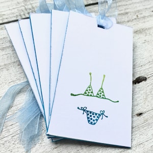 May include: A stack of blank gift tags with blue trim and a blue ribbon. The tags feature a hand-drawn illustration of a green and blue polka dot bikini.