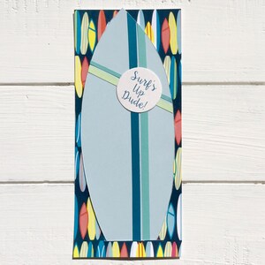 Op de afbeelding: Een blauw en wit surfplank met een gele streep en een ronde sticker met de tekst "Surf's Up Dude!" De achtergrond is een patroon van kleurrijke surfplanken.